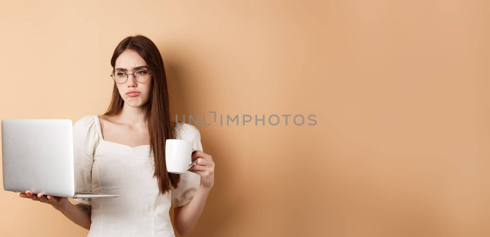 Tired working girl looking at laptop bored, drinking coffee while using computer for work, standing on beige background.