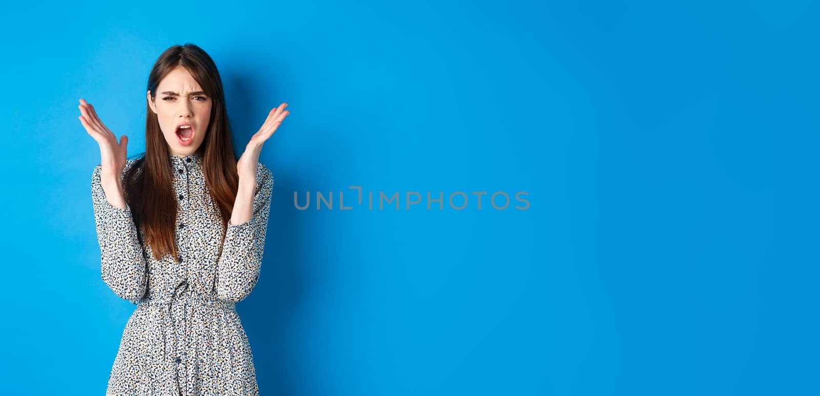 Frustrated and angry woman in dress, scream and frown at camera, raising hands up outraged, having an argument, standing on blue background by Benzoix