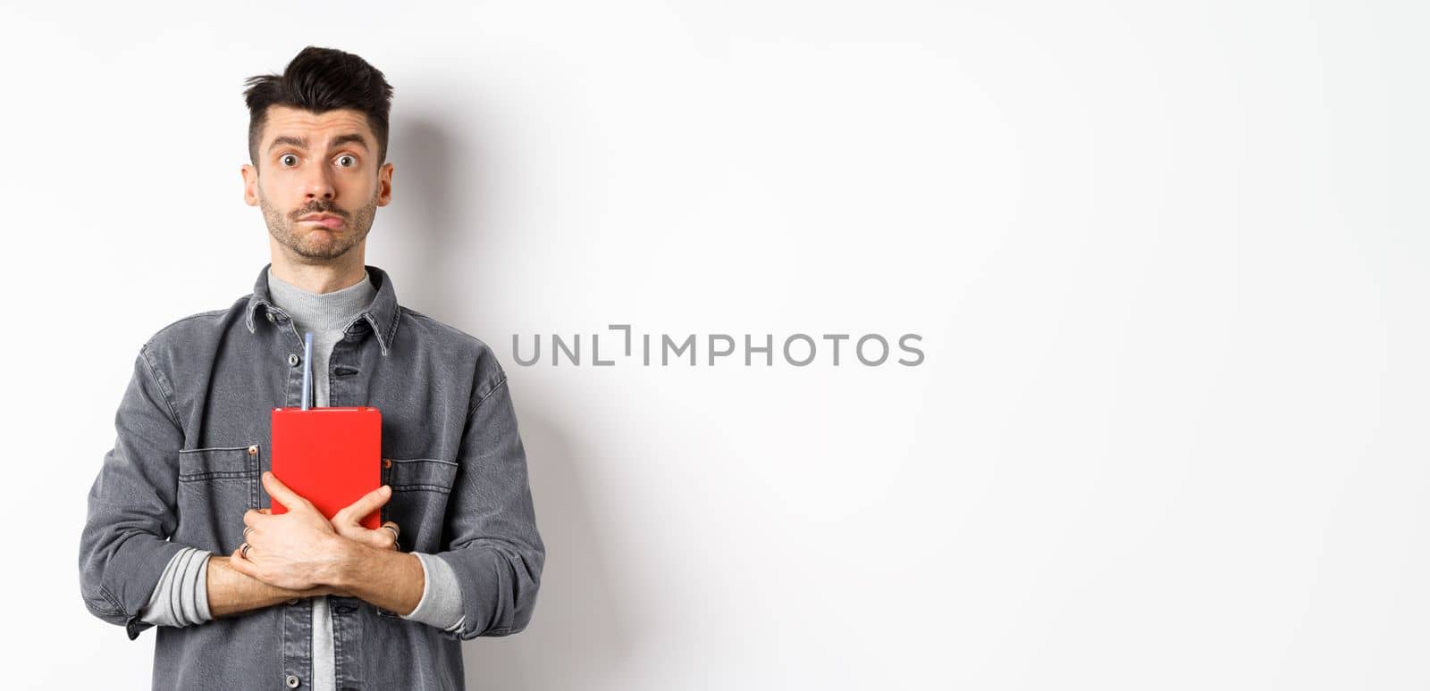Cute and shy guy hugging his diary, holding red journal and look modest at camera, standing against white background by Benzoix