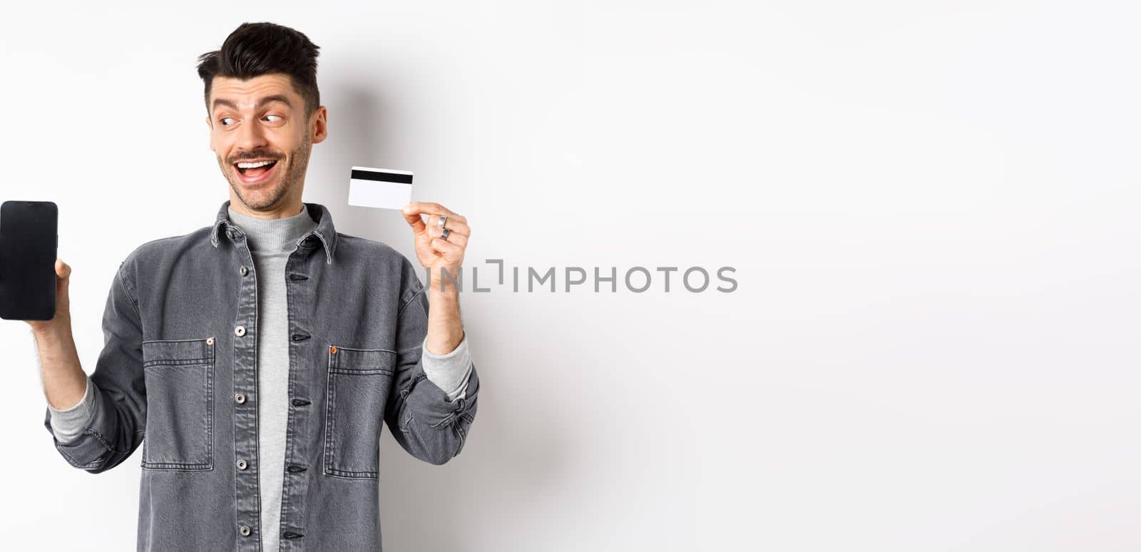 Handsome smiling man in denim jacket looking at empty smartphone screen and showing plastic credit card, good internet offer, standing against white background.