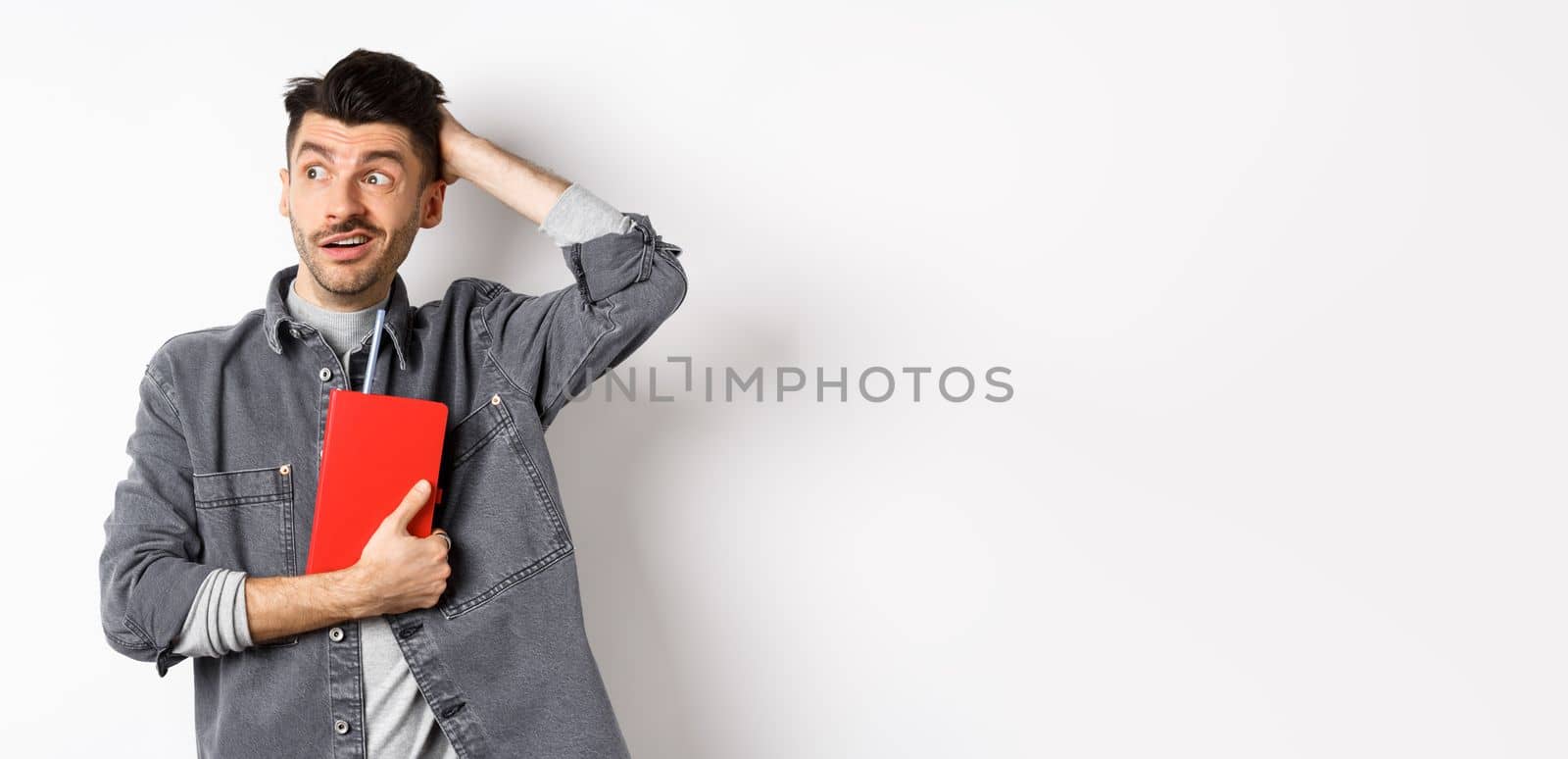 Confused guy scratch head and look aside at logo puzzled, holding red diary or planner, standing against white background by Benzoix