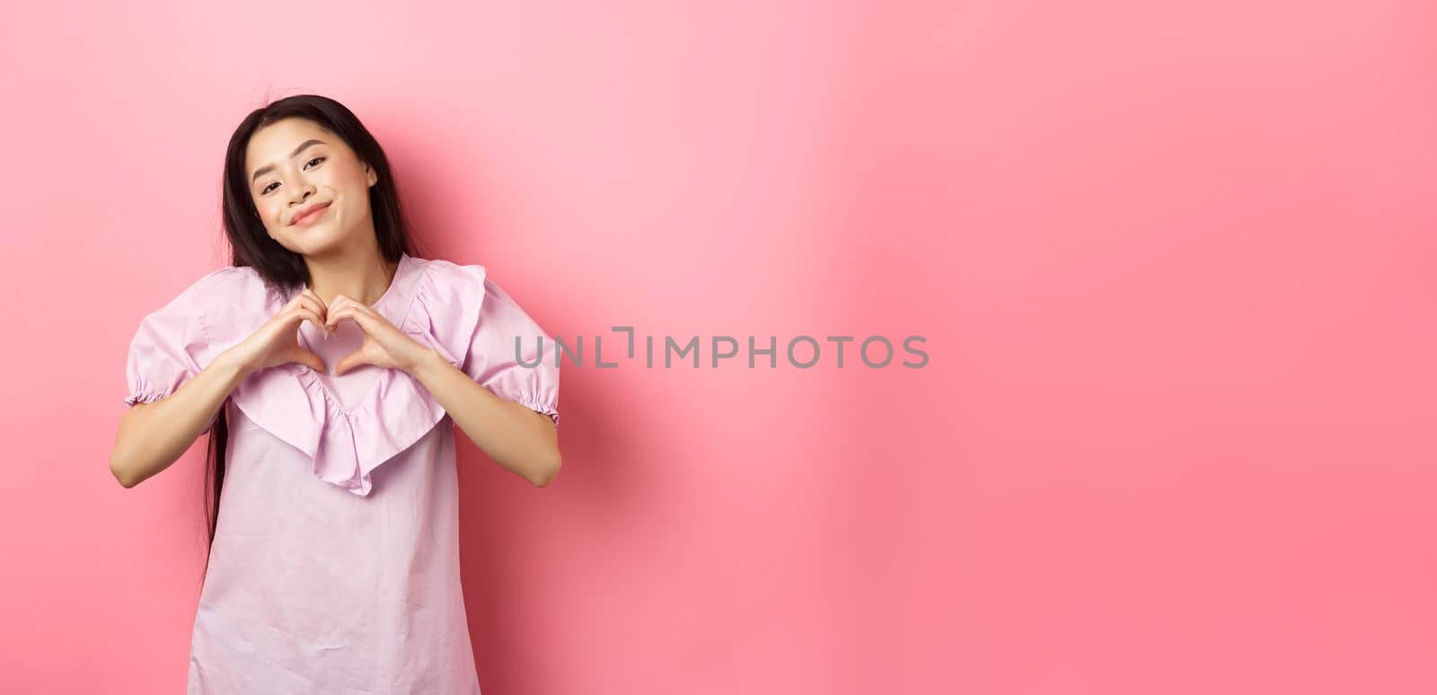 Valentines day concept. Beautiful korean teenager in dress showing heart gesture and smiling with admiration and affection, falling in love, feel romantic, pink background.