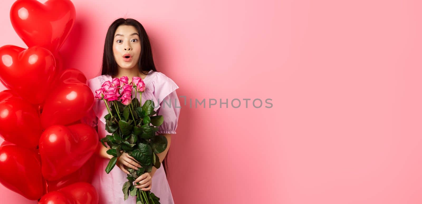 Surprised asian girl in dress standing near valentines day heart balloons and say wow at camera, holding flowers bouquet from lover, romantic date with roses, pink background by Benzoix