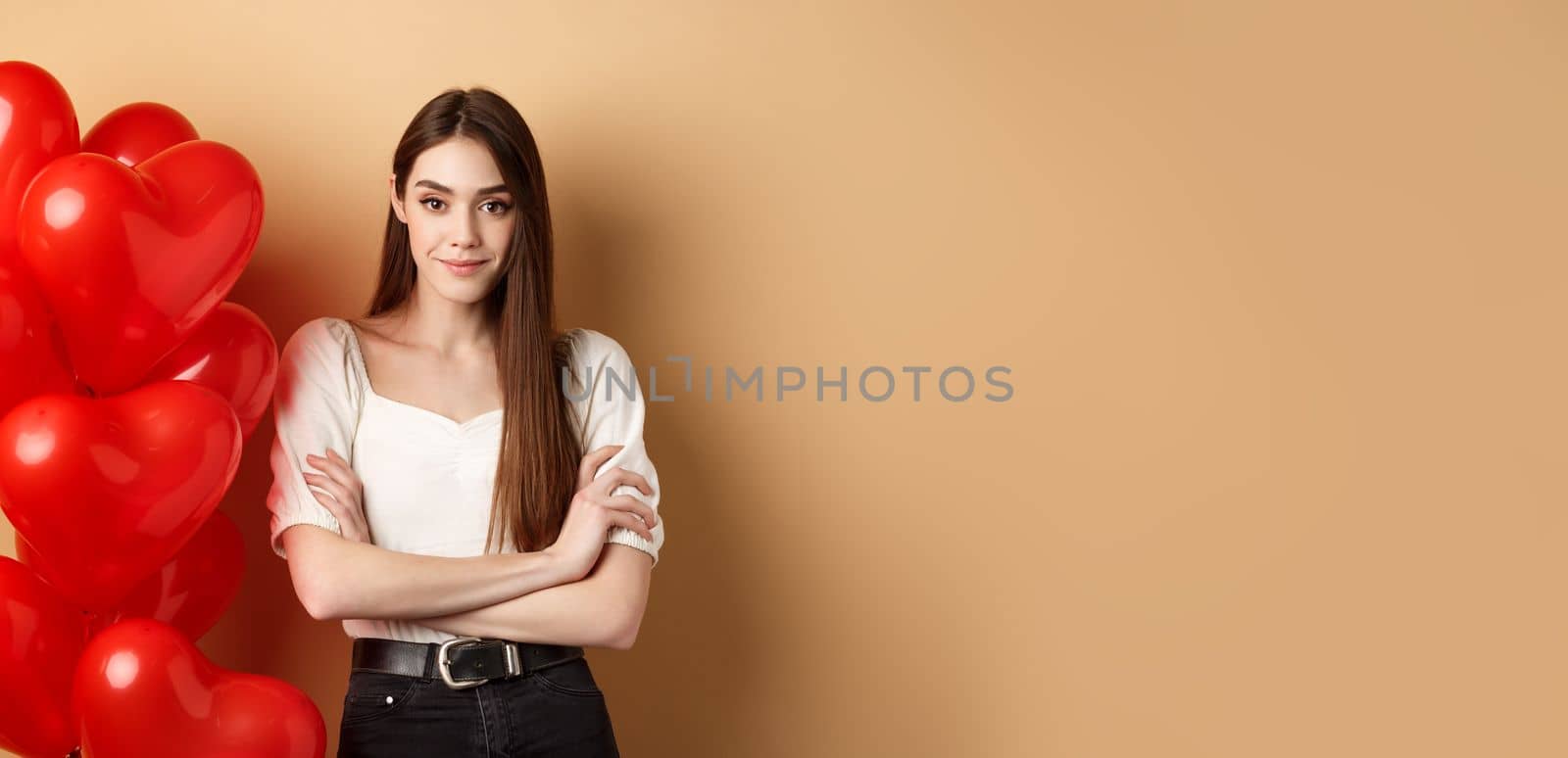 Sassy young girl cross arms on chest and smiling confident, standing near Valentines day hearts balloons and looking at camera, beige background.