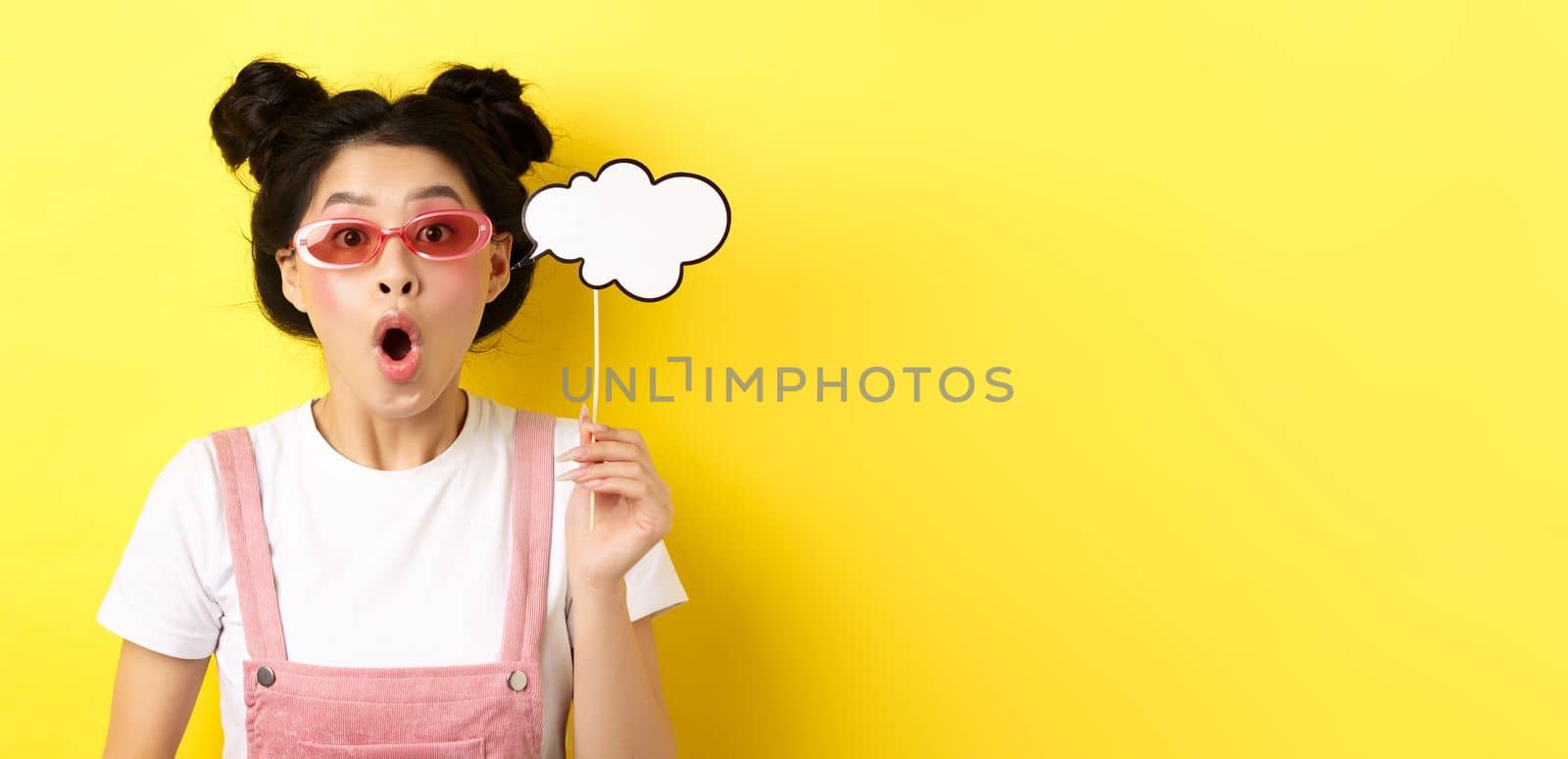 Summer and fashion concept. Excited party girl in sunglasses, holding comment cloud party mask and gasping amazed, standing impressed on yellow background by Benzoix