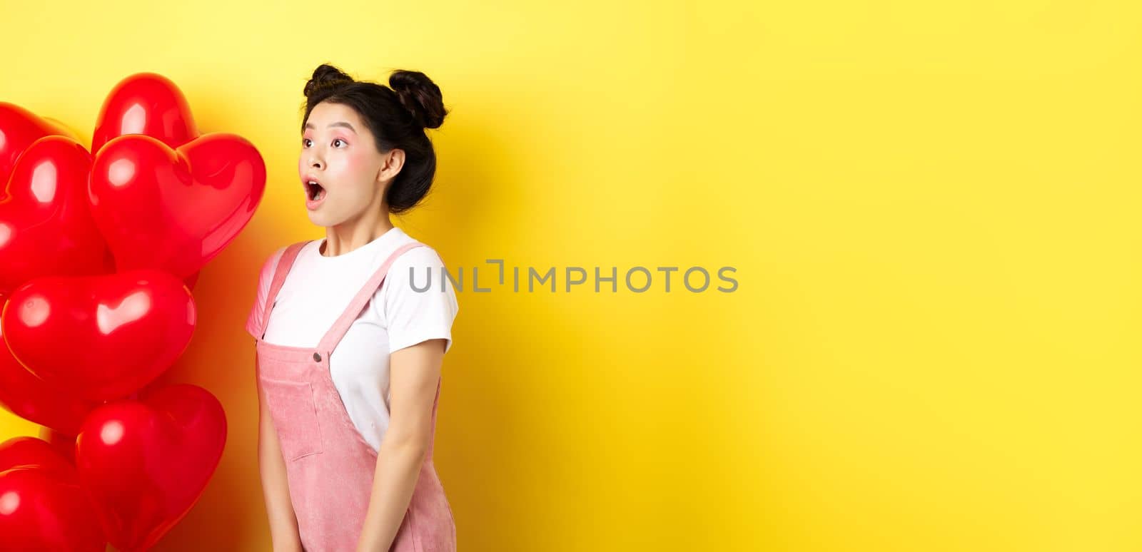 Valentines day and relationship concept. Fashion girl looking amazed, gasping and say wow, staring left while standing near red hearts balloons, surprise from lover, yellow background by Benzoix