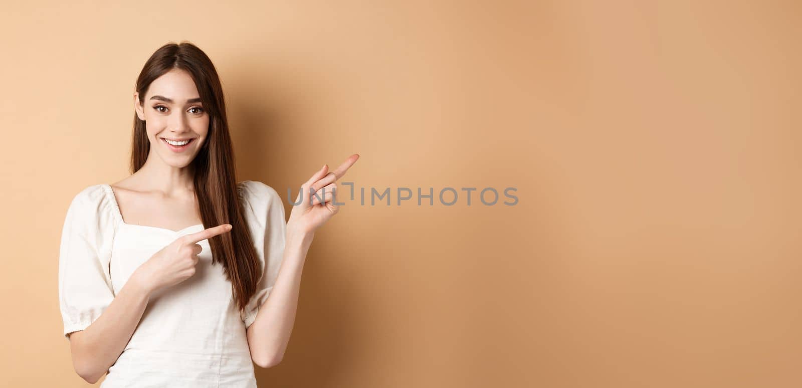 Smiling young woman in dress pointing aside and looking satisfied, showing good promo deal, standing on beige background.