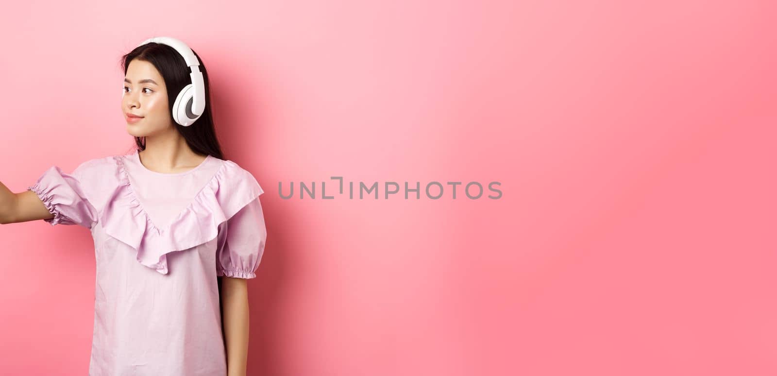 Beautiful japanese girl taking selfie on smartphone, wearing wireless headphones, standing against pink background by Benzoix