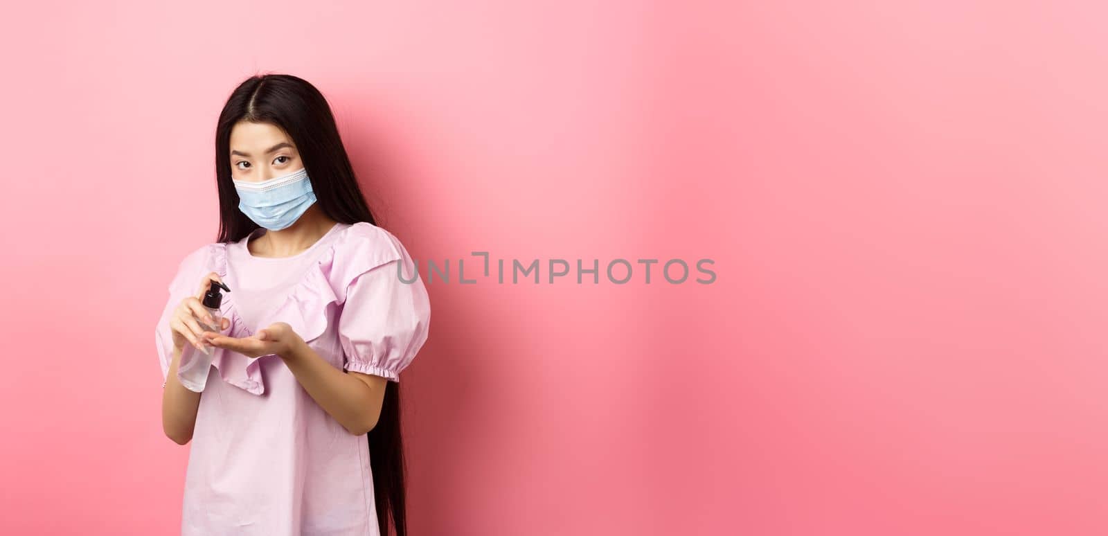 Covid-19, lifestyle and health concept. Beautiful asian woman in medical mask clean hands with sanitizer, apply antiseptic on palms, standing on pink background.
