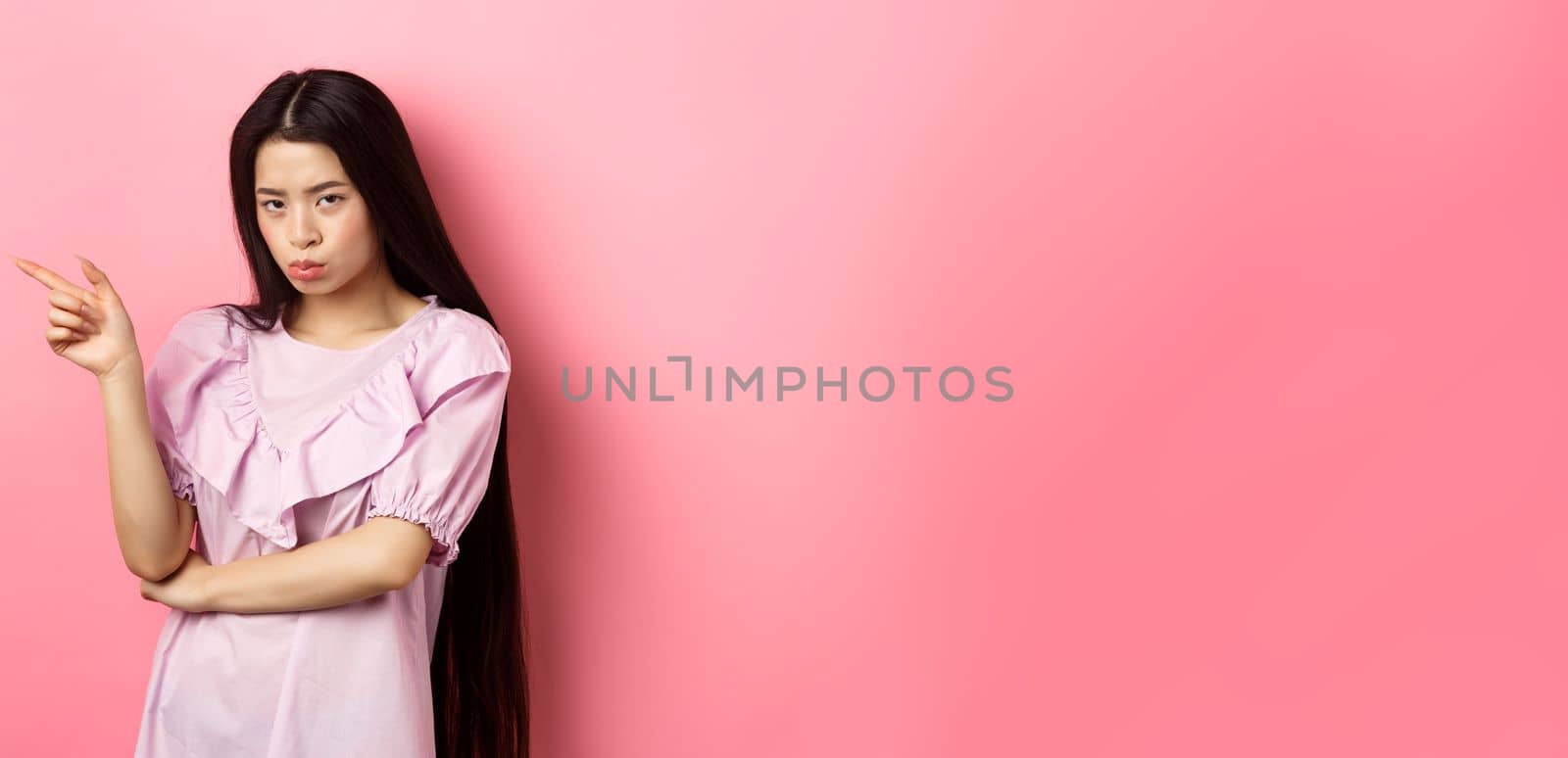 Sad and grumpy asian girl sulking, pointing left and look at camera jealous, want something, complaining and showing desired thing, standing on pink background by Benzoix
