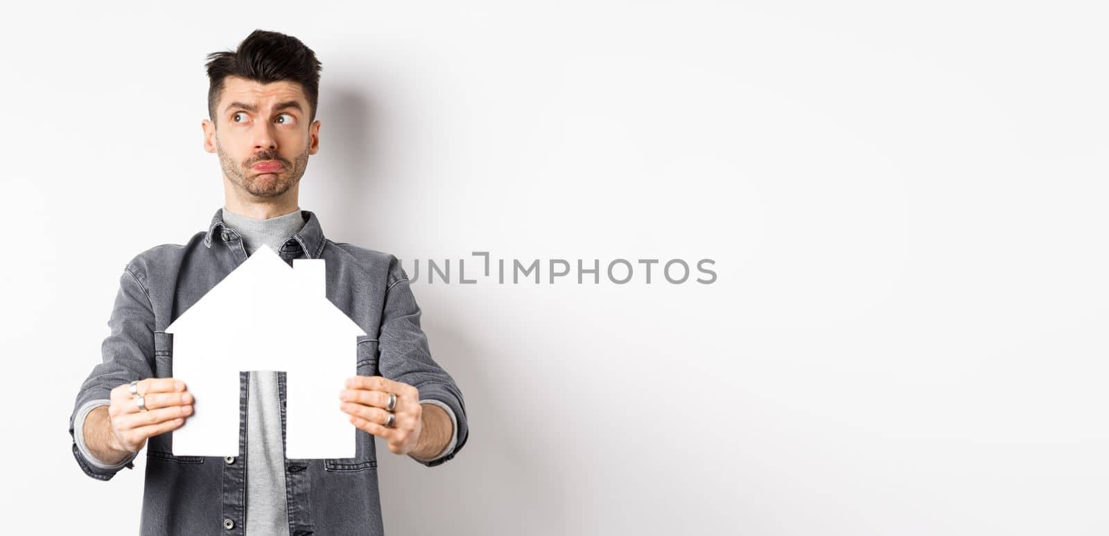 Real estate and insurance concept. Indecisive guy look aside pensive and showing paper house cutout, thinking of buying property, standing against white background.