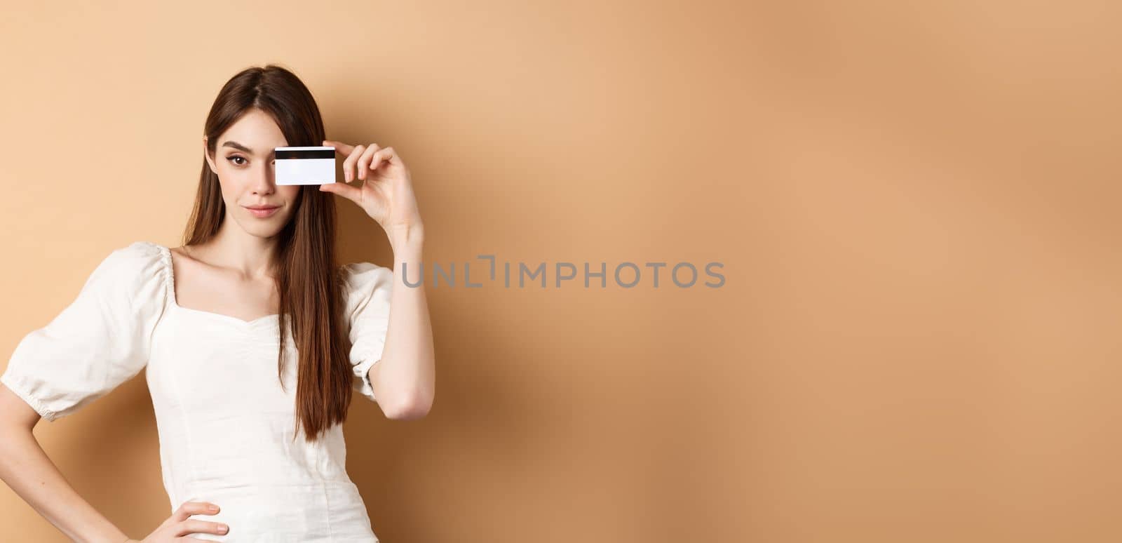 Sassy young woman showing plastic credit card on face and looking determined, going on shopping, standing against beige background.