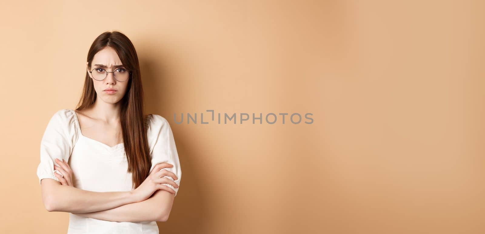 Angry and offended girl in glasses frowning, cross arms on chest and sulking, standing defensive on beige background.
