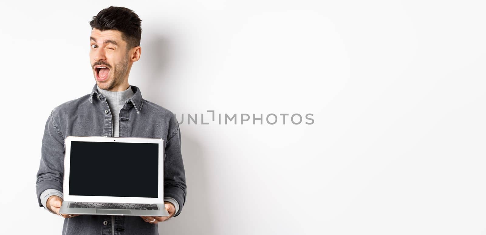 Cheerful guy winking and showing empty smartphone screen, offer you advertisement, standing on white background by Benzoix