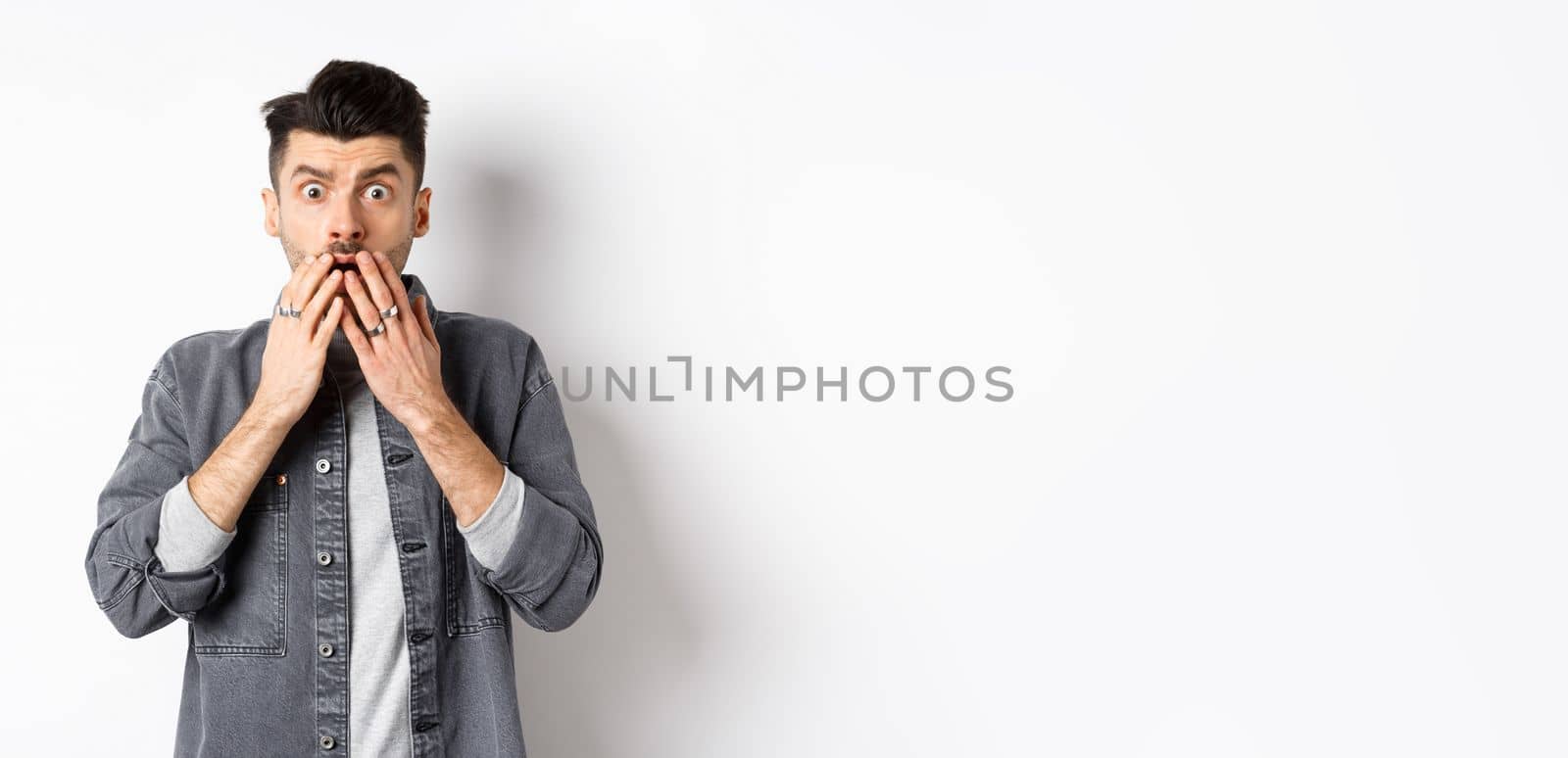 Shocked caucasian guy gasping alarmed, cover mouth with hands and staring at something terrible, witness bad accident, say oh my god, standing on white background.