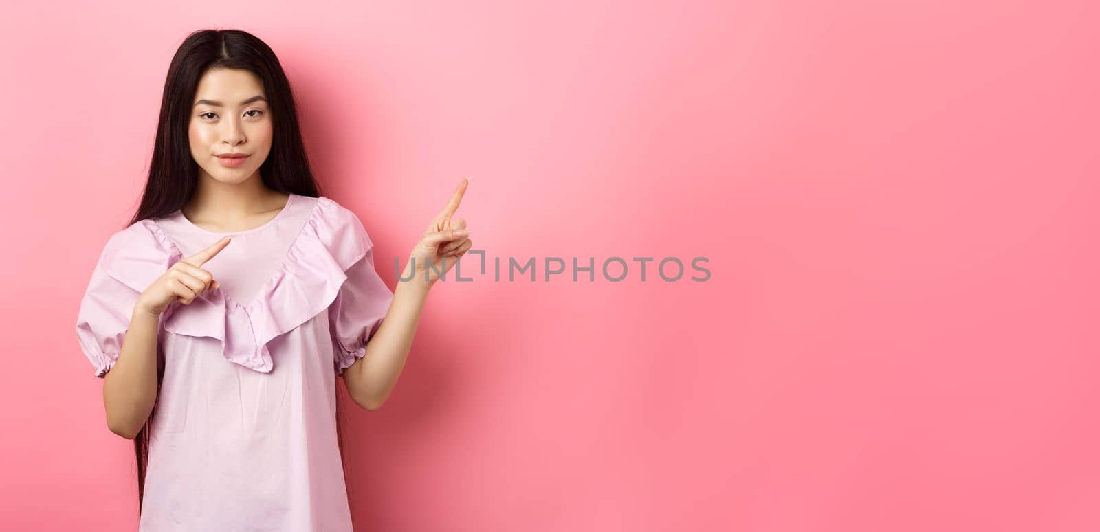 Cheeky asian girl showing interesting advertisement, pointing fingers right at logo and smiling cunning, hinting on good deal, pink background.