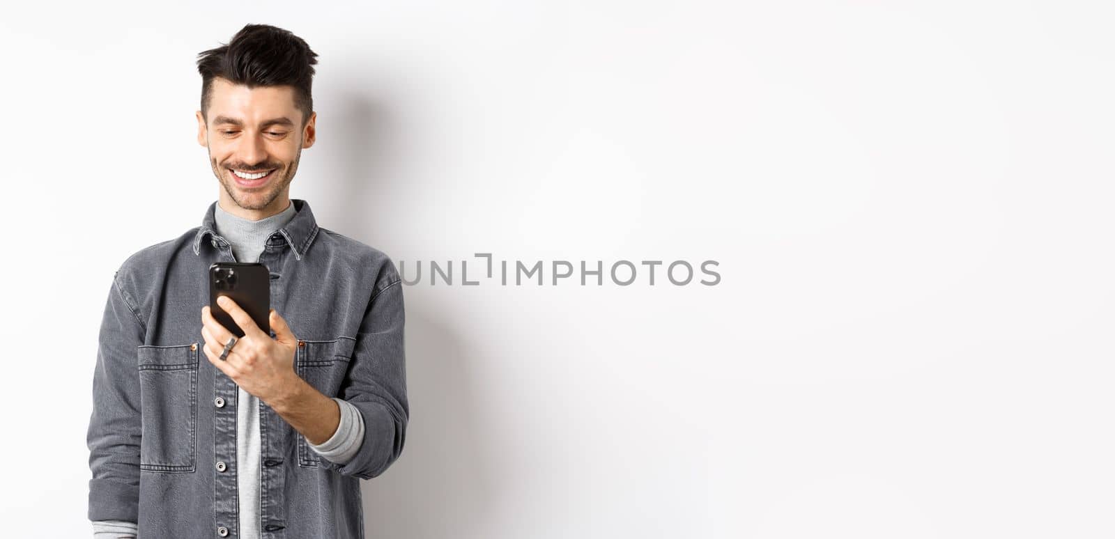 Handsome man reading message on phone and smiling. Guy holding smartphone and looking at screen, standing on white background.