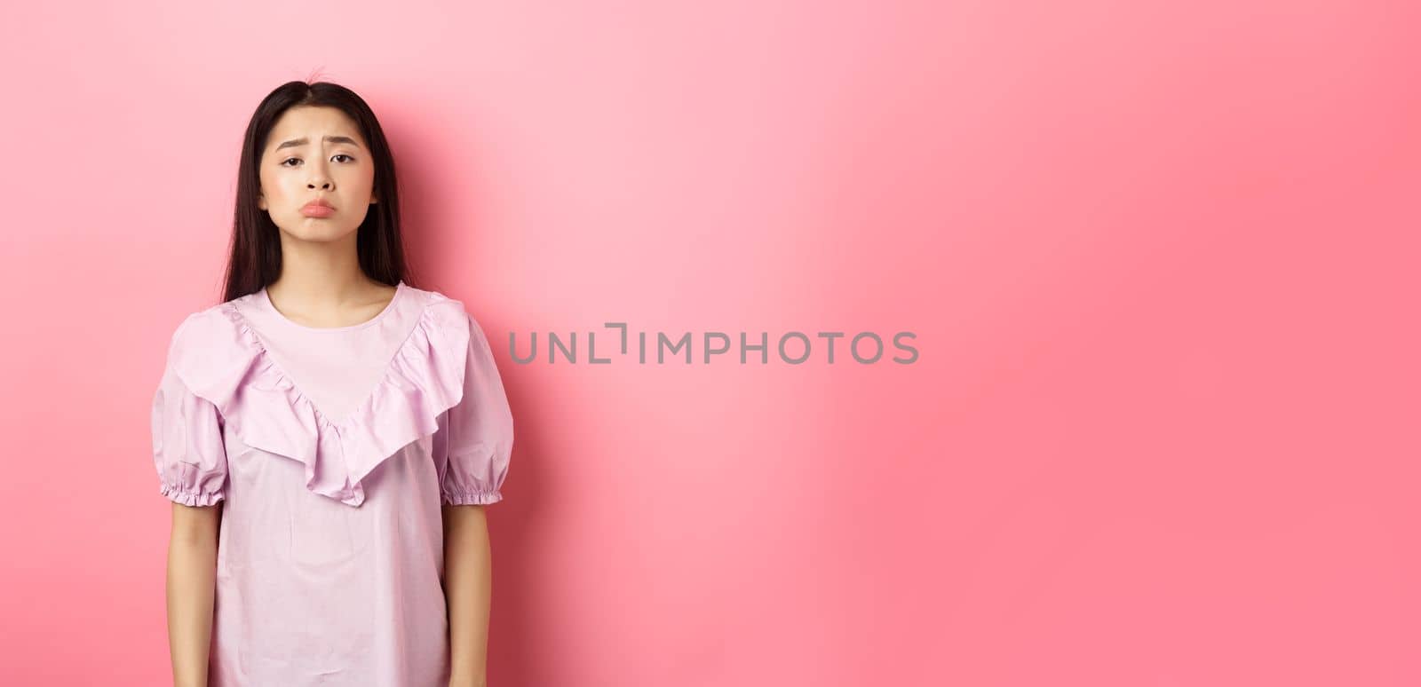 Sad whining asian girl pouting and frowning, looking upset about something unfair, complaining, standing in dress against pink background.