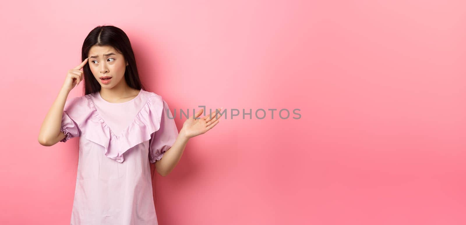 Are you stupid. Shocked asian woman pointing at head and complaining, scolding someone crazy or strange, look aside, standing against pink background.