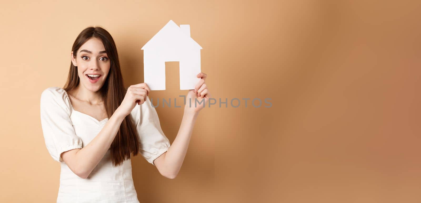 Real estate. Excited woman got new apartment, showing paper house cutout and smiling happy, standing on beige background.