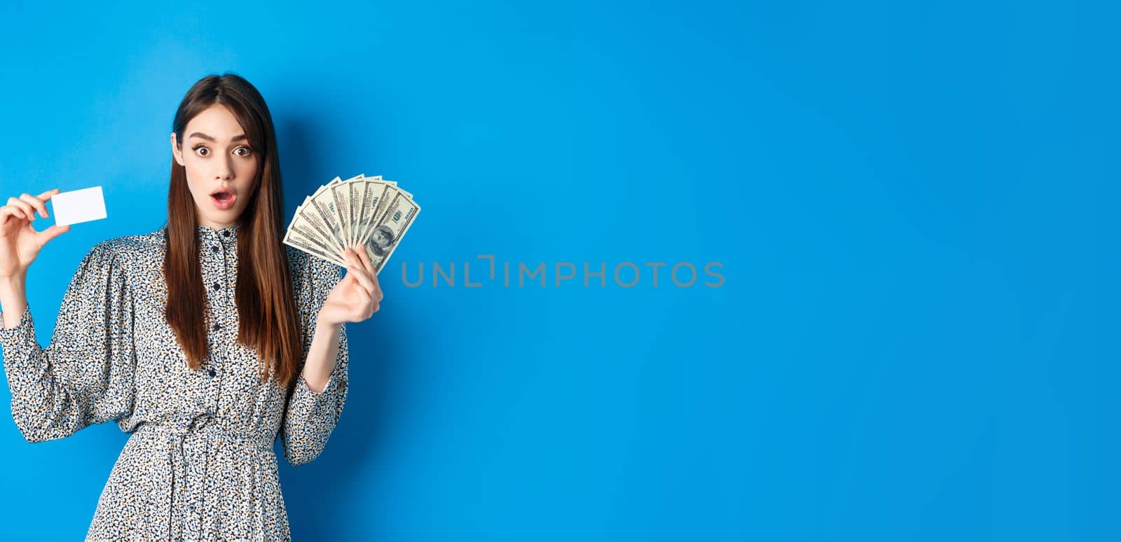 Excited lucky girl showing plastic credit card and money dollar, gasping amazed, buying something expensive, standing on blue background.