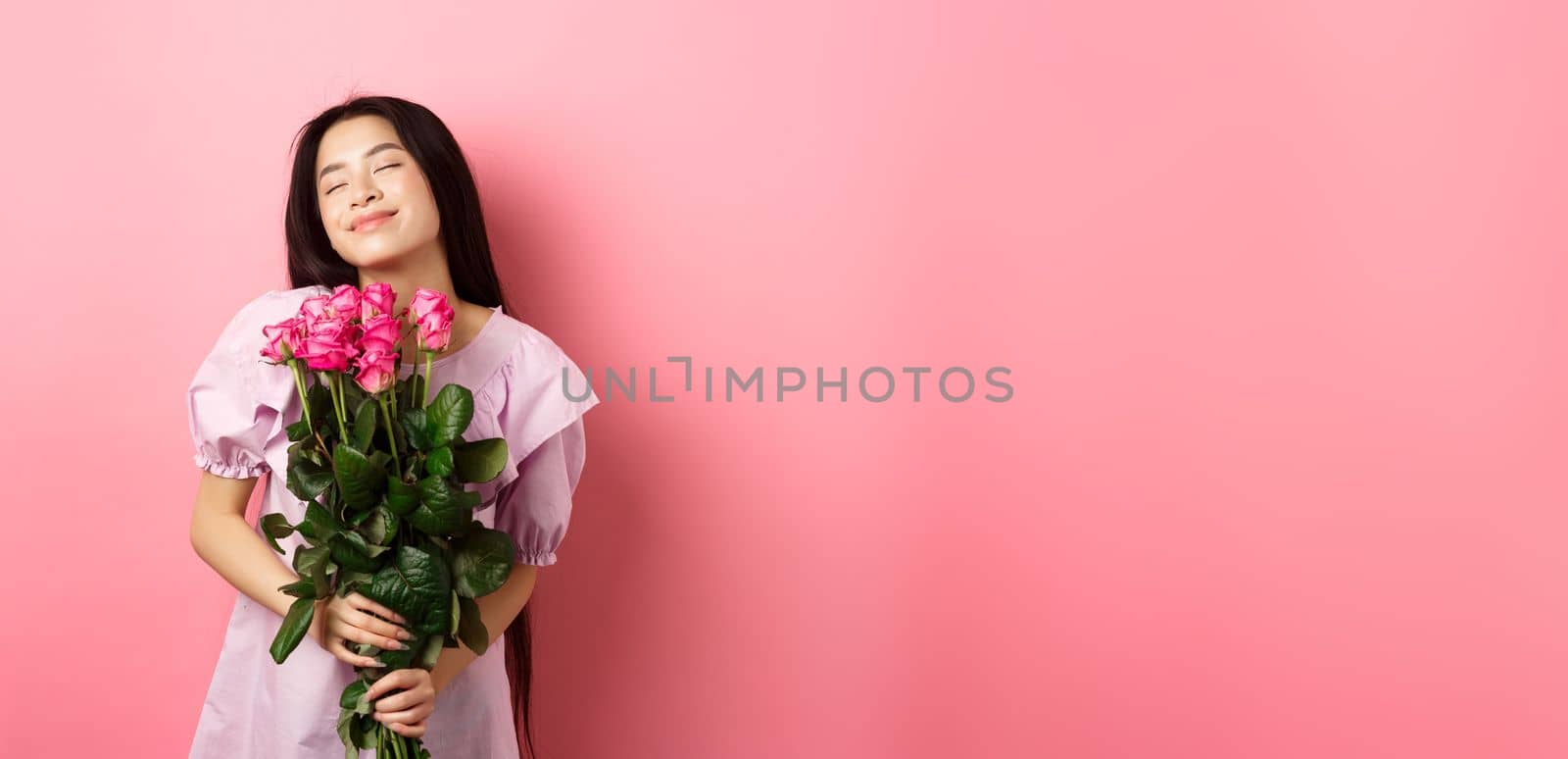 Dreamy asian teenage girl feeling romantic, holding flowers and dreaming, imaging valentines day date, wearing cute dress, standing on pink background.