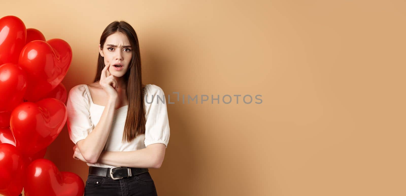 Valentines day and love concept. Confused and shocked girl staring displeased at camera, standing near romantic red hearts balloons on beige background.