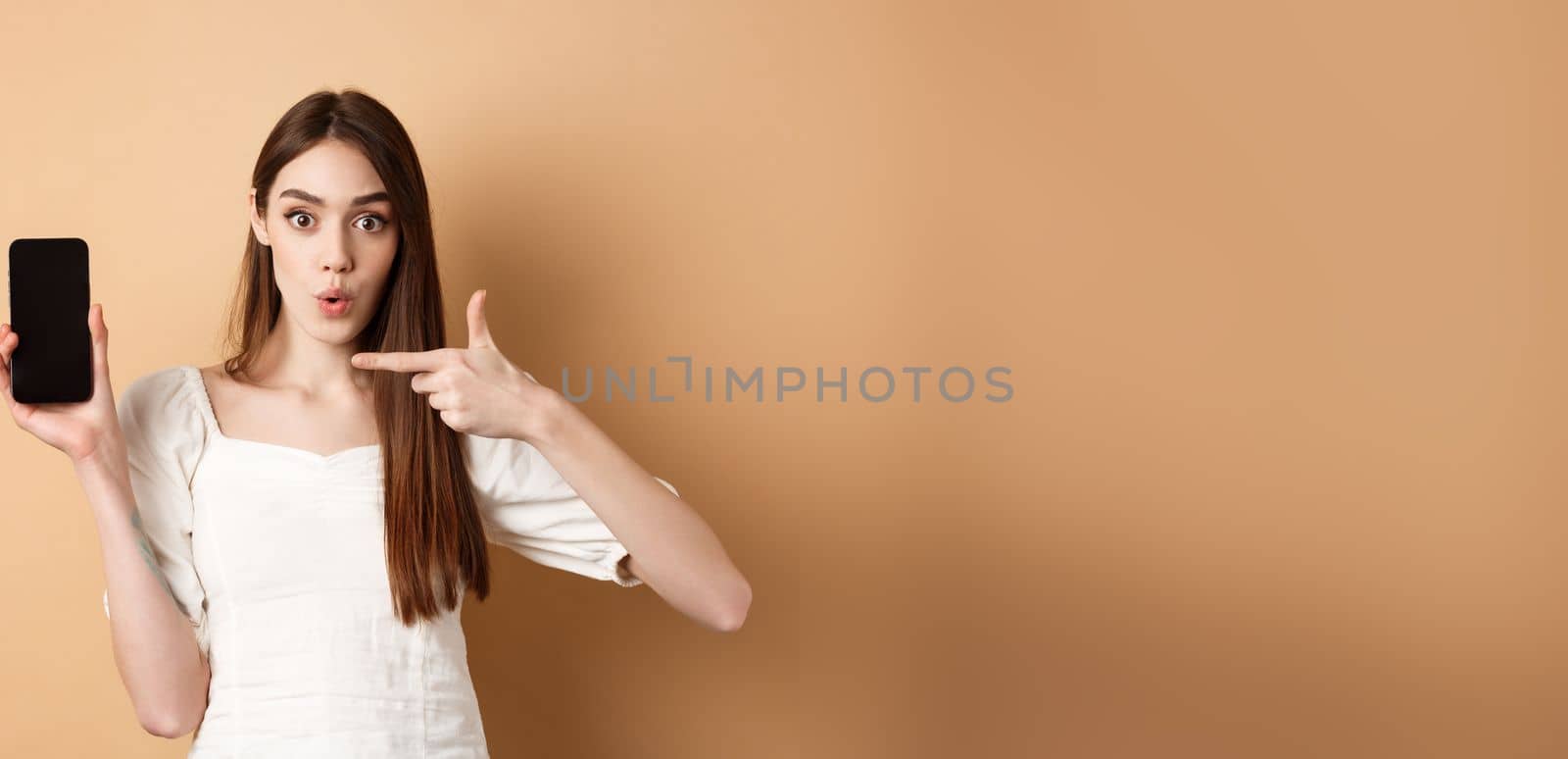 Excited woman showing news on screen, pointing at empty phone and looking surprised, standing on beige background.