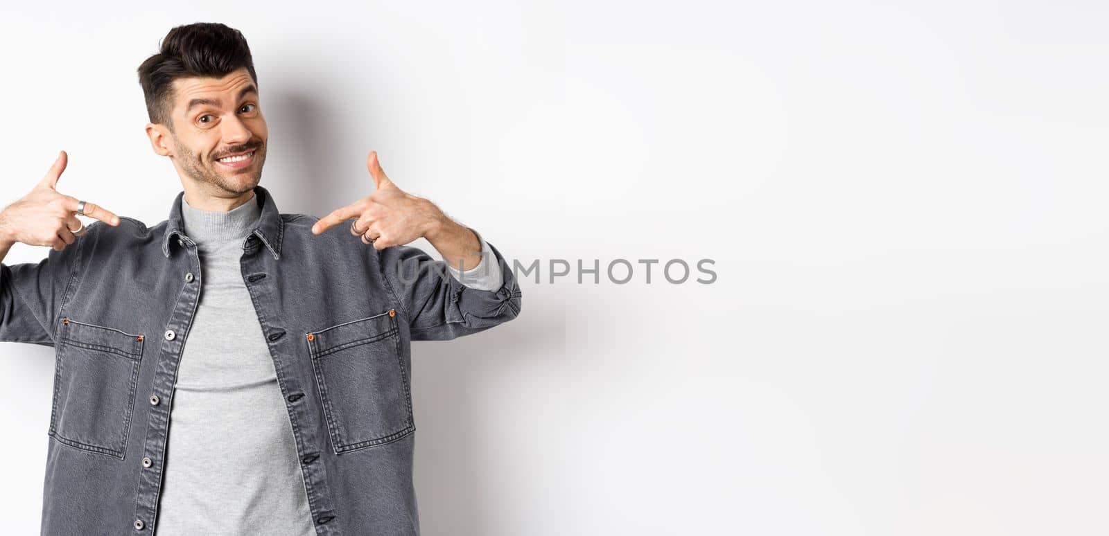 Handsome guy with moustache and white smile pointing fingers at center, showing company logo, standing on white background.