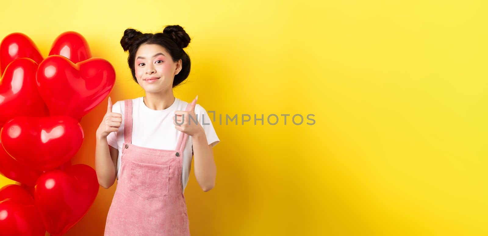 Cute teenage asian girl showing thumbs up, waiting for Valentines day near red heart balloons, wearing outfit for romantic date, yellow background by Benzoix