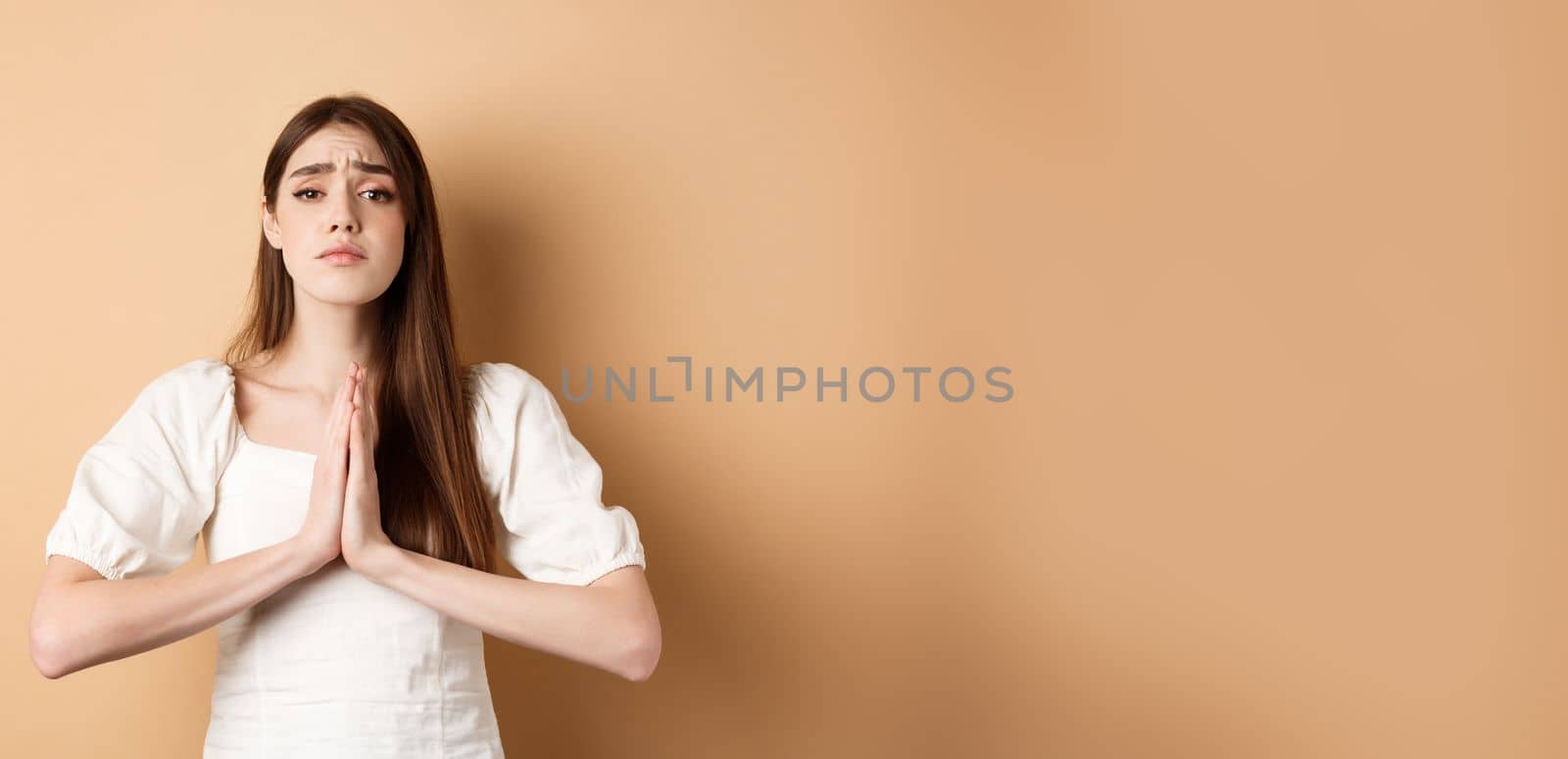 Please help. Desperate young woman begging you, holding hands in pleading gesture and looking sad at camera, asking for favour, beige background.
