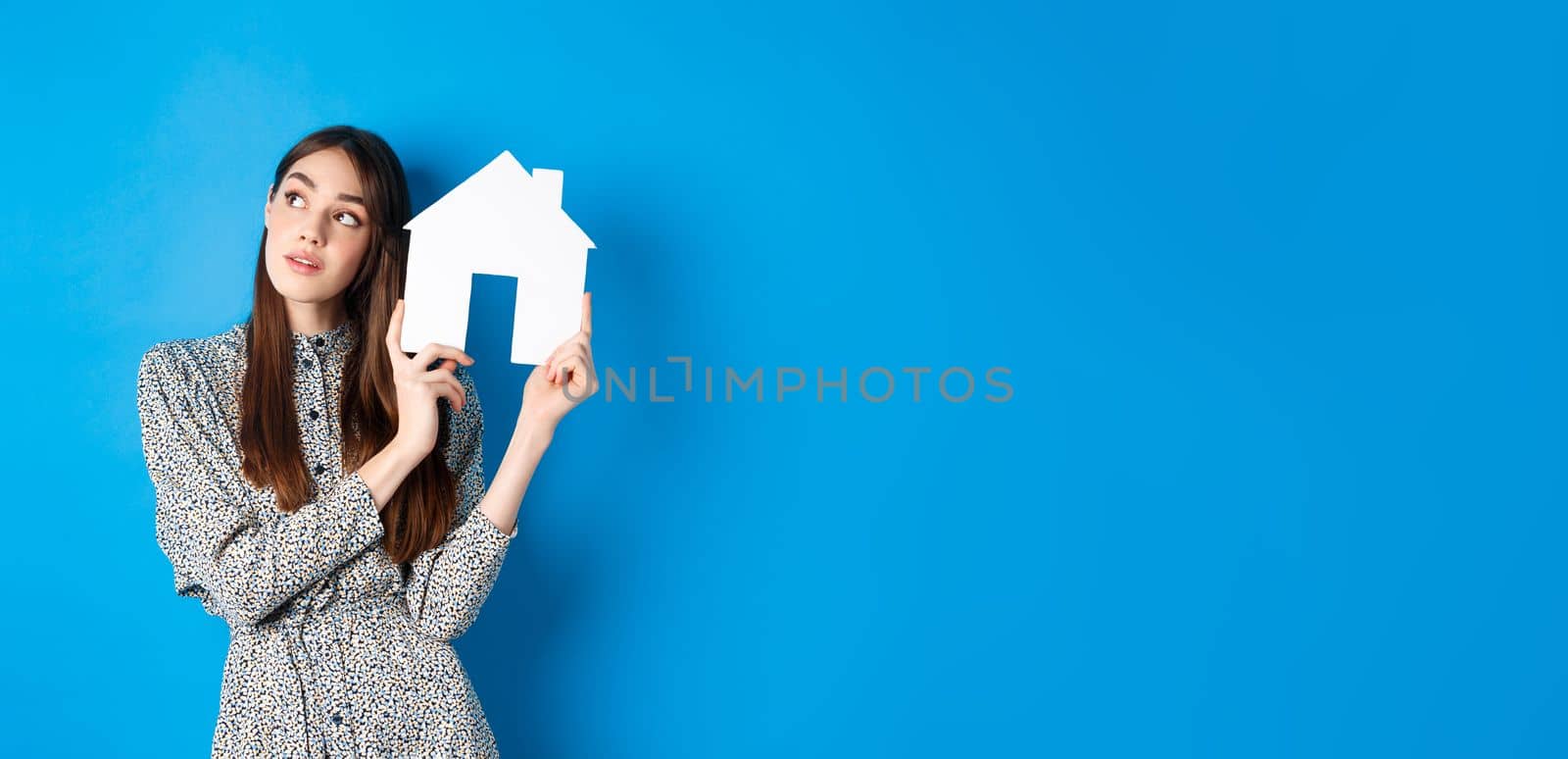Real estate, realtors and insurance concept. Dreamy beautiful adult woman thinking of own house, showing paper home cutout and look aside at logo, standing against blue background.
