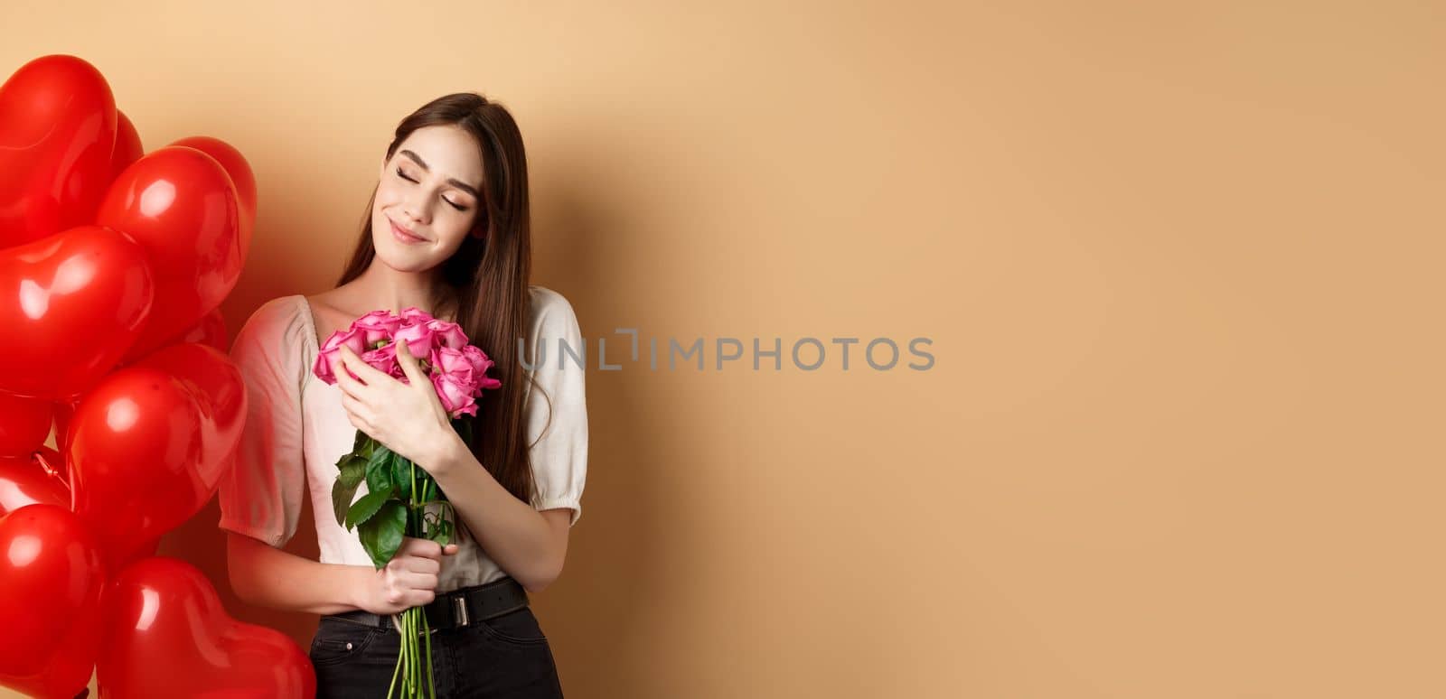 Sensual and romantic girl hugging bouquet of pink roses, smiling with eyes closed, thinking of lover, standing near Valentines day heart balloons, beige background.