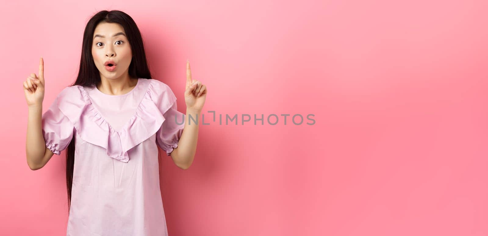 Excited asian girl showing awesome promo offer, pointing fingers up and gasping amazed, standing on pink background.