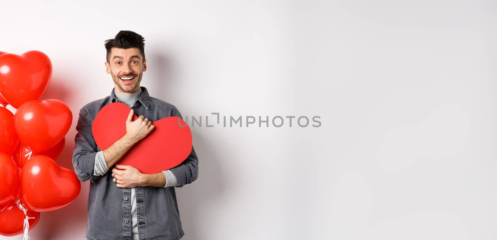 Young romantic man looking with love at camera, hugging Valentine heart card and smiling happy, celebrating lovers holiday, standing near red balloons, white background by Benzoix