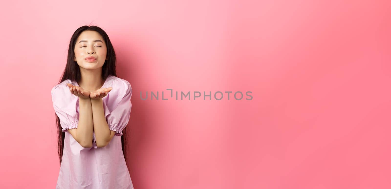 Cute and romantic asian girl blowing air kiss at camera, close eyes and smile tenderly, standing in dress against pink background.