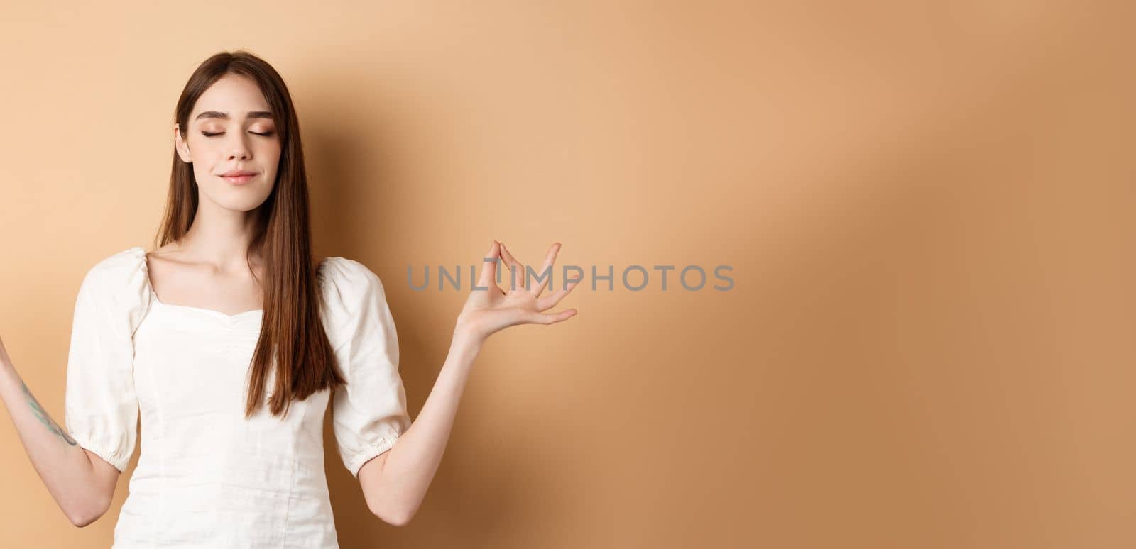 Relaxed young woman meditating, holding hands in yoga zen pose and close eyes, feel peace and smiling, standing on beige background by Benzoix