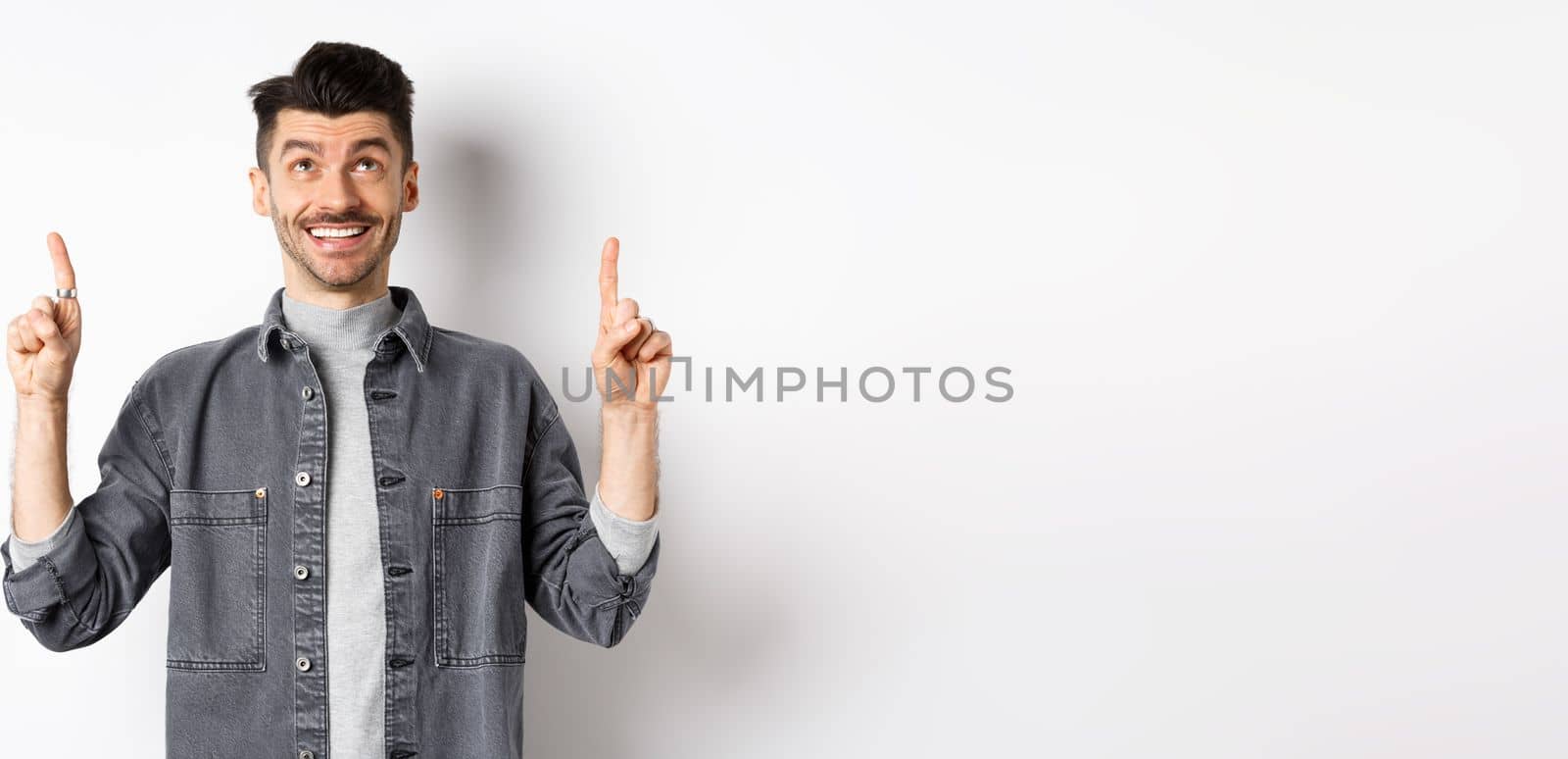 Cheerful caucaisan man with moustache pointing and looking up, smiling dreamy, showing cool promotion deal, standing on white background.