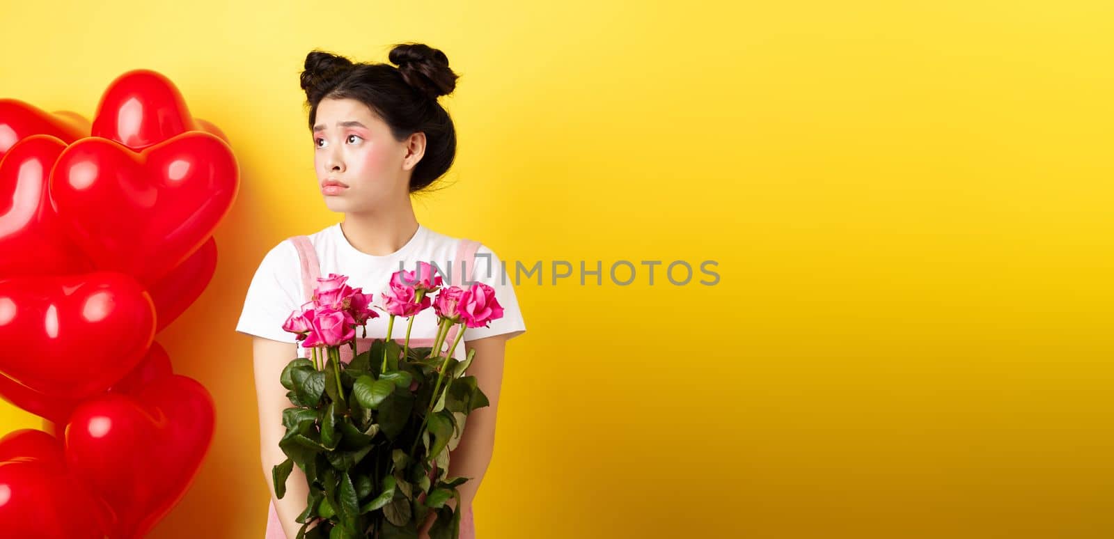 Happy Valentines day. Sad and lonely girl looking left upset, holding bouquet of roses, standing alone near red hearts balloons and frowning, yellow background.