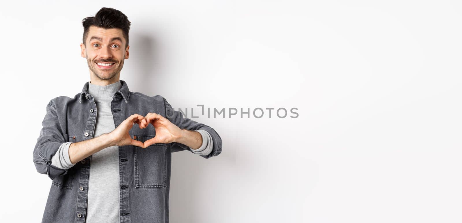 Smiling boyfriend showing heart with love, looking romantic and happy at lover, standing on white background. Valentines day and relationship concept by Benzoix
