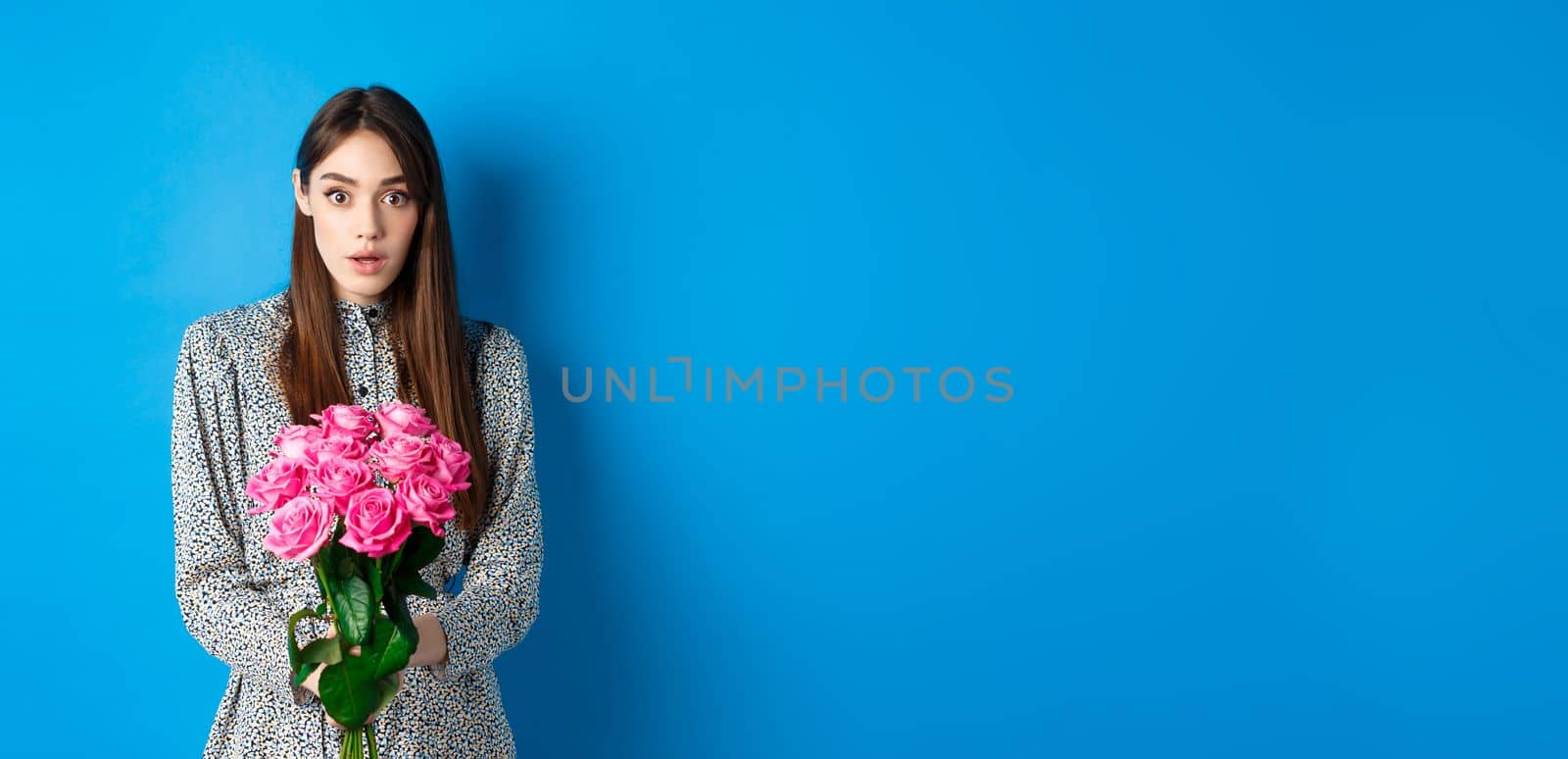 Valentines day concept. Surprised girlfriend receiving beautiful bouquet of flowers and looking with disbelief at camera, standing on blue background.