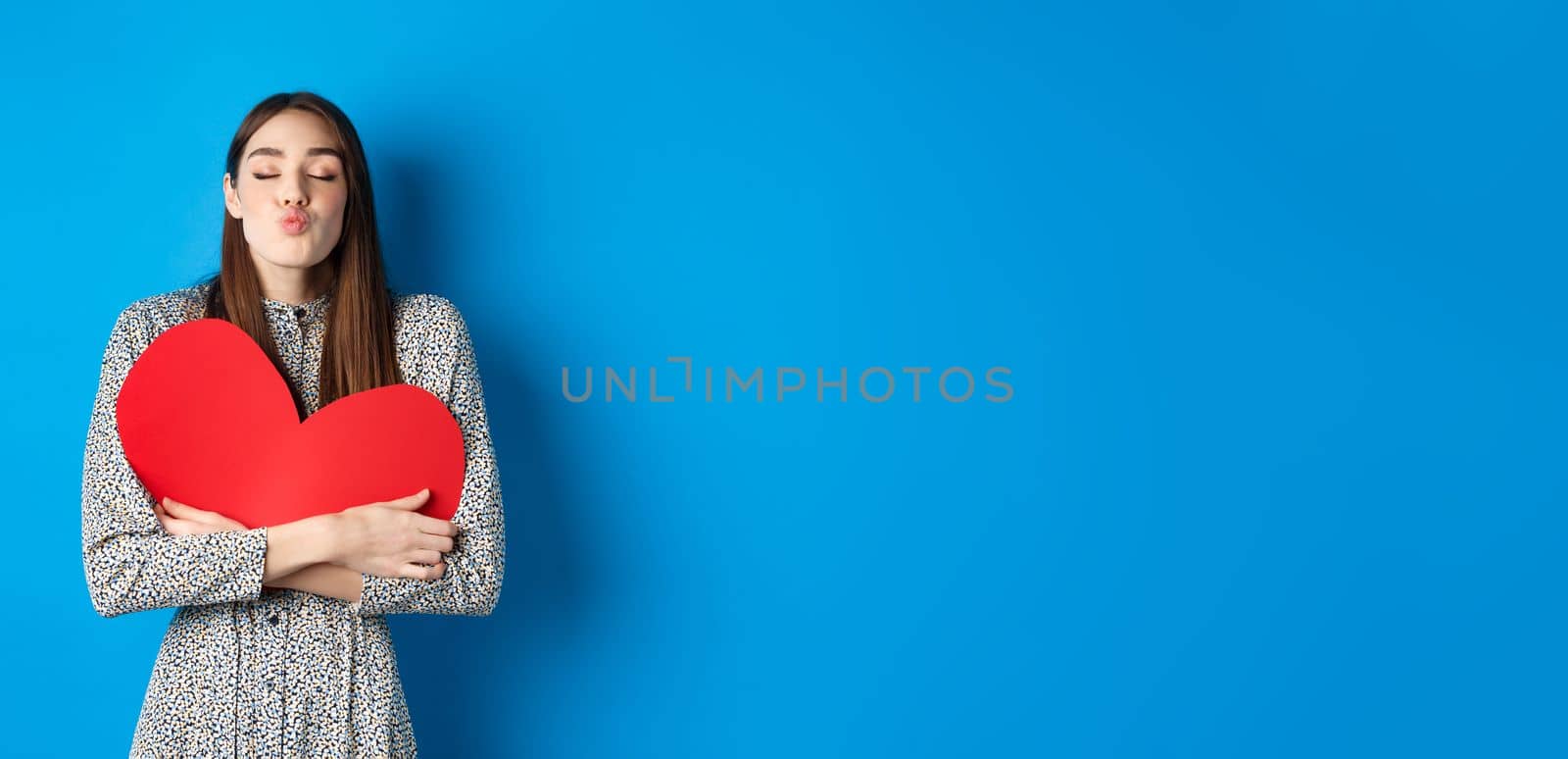 Valentines day. Romantic beautiful woman close eyes and pucker lips for kiss, holding big red heart cutout, kissing you, standing on blue background.