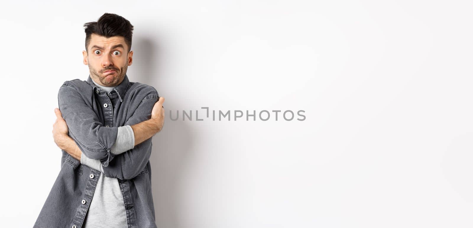 Funny young man hugging himself and grimacing, feeling embarrassed and exposed, covering body from people glances, white background.