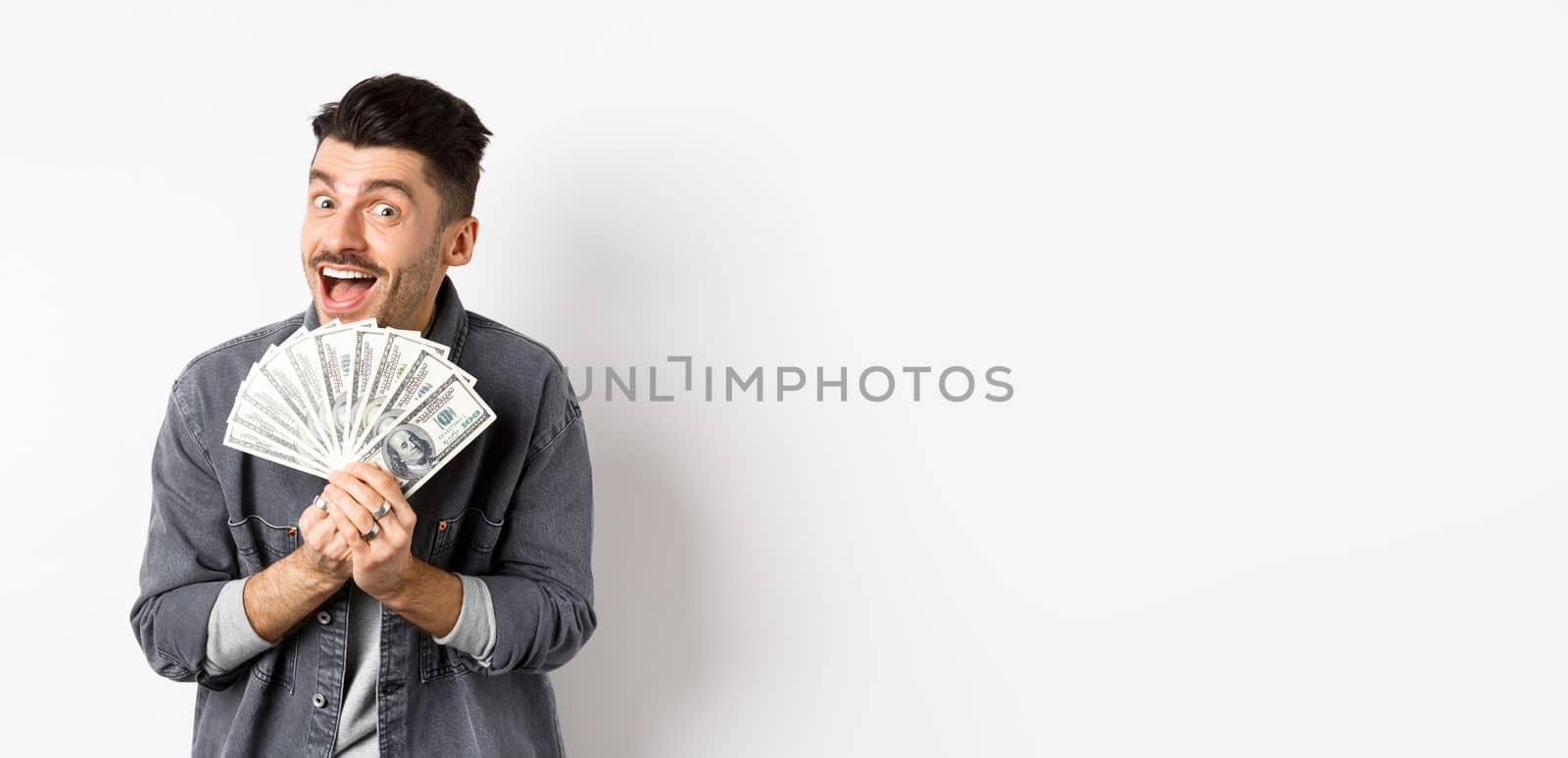 Excited young man smiling and showing dollar bills, making money, standing against white background.