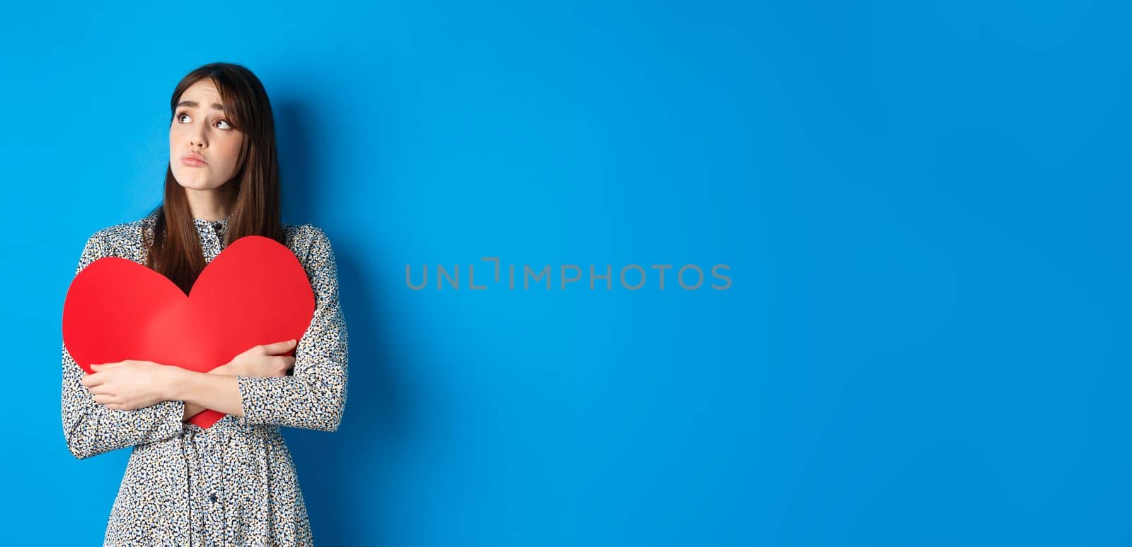 Valentines day. Sad heartbroken girl sobbing, looking aside at empty space and crying upset, hugging big red heart, feeling lonely without lover, standing alone on blue background by Benzoix