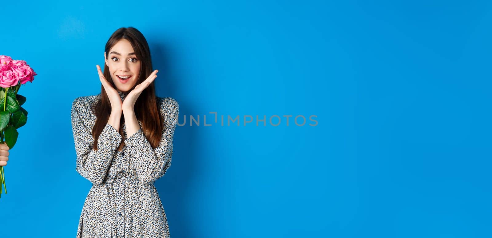 Valentines day concept. Excited and happy young woman looking amazed at camera while hand stretch out hand with bouquet of flowers, receiving romantic gift, blue background.