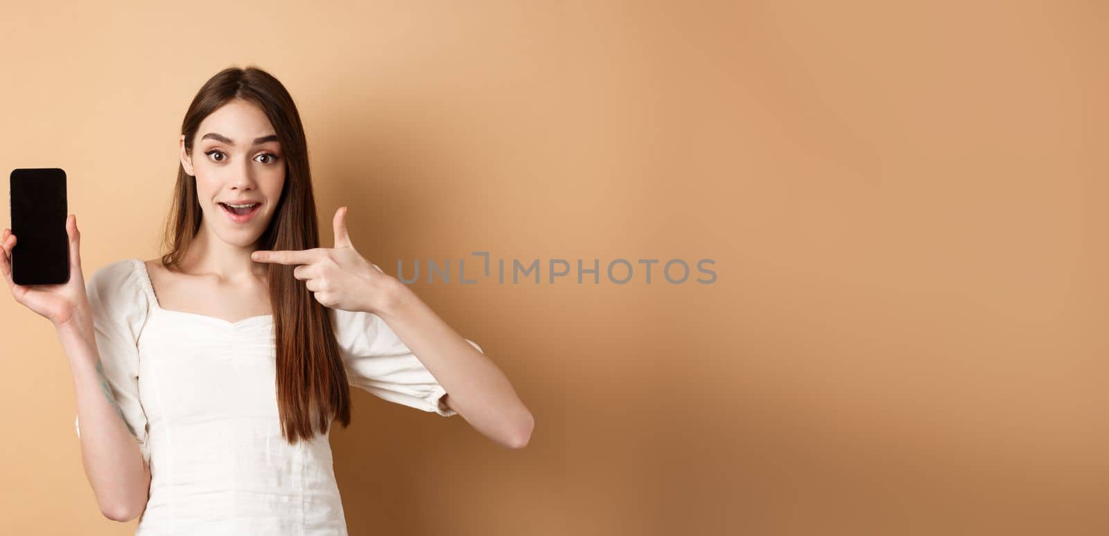 Cheerful woman pointing finger at empty phone screen, looking excited, standing on beige background.