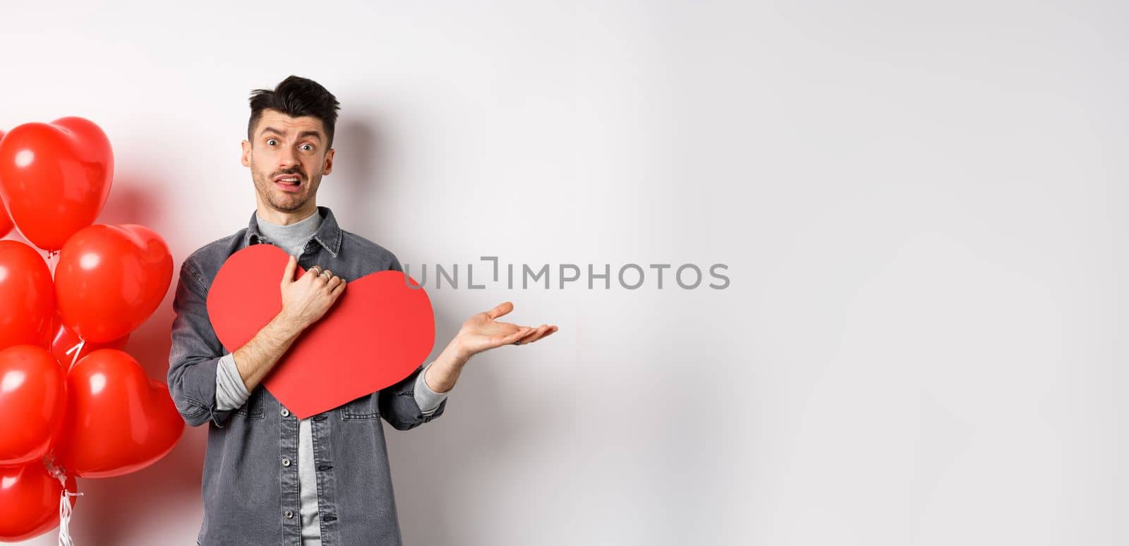 Confused man hugging big Valentine card and shrugging, feeling careless about romantic holiday, standing skeptical on white background.