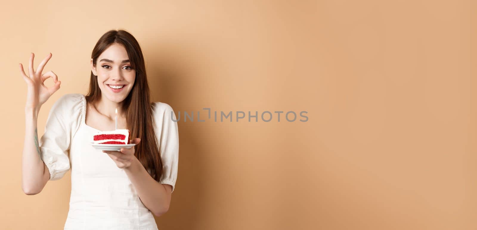 Happy birthday girl show okay gesture and hold bday cake, making wish on her holiday, standing on beige background.