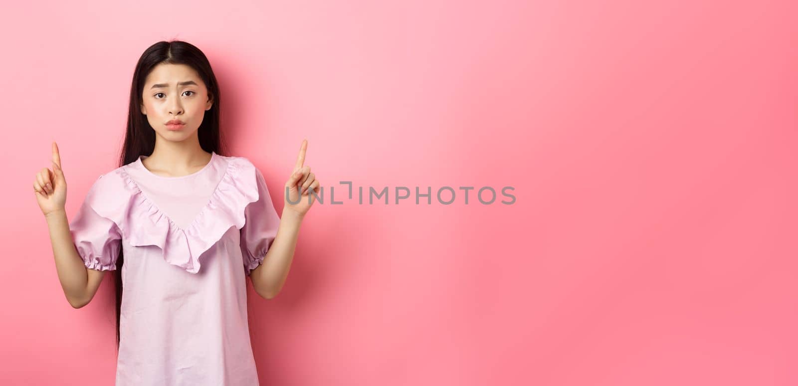 Cute timid asian woman pointing fingers up, frowning and look upset, pointing fingers up at logo, standing in dress on pink background.